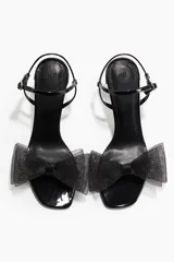 Sandalias de taco alto negro con acabado charolado, tira delantera con detalle de lazo voluminoso de tul y tira ajustable al tobillo con hebilla metálica.