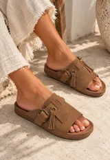 Sandalias planas color beige, fabricadas en gamuza, con tira ancha en el empeine, hebilla metálica de ajuste y detalle de flecos colgantes con argolla.