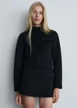 Chaqueta corta de diseño recto, color negro, con cuello de tira, cierre frontal con cremallera, bolsillos laterales y un bolsillo de ribete en el pecho.