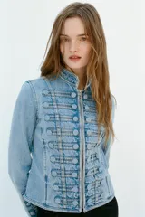 Chaqueta denim entallada de cuello redondo y manga larga, con cierre frontal de cremallera metálica. Presenta detalles de aplicaciones en el mismo tejido y pasamanería frontal de inspiración militar.