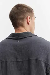 Camisa de manga corta color beige confeccionada en muselina de algodón, con cuello abierto, cierre frontal de botones y un bolsillo aplicado en el pecho.