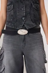 Cinturón de cuero negro con hebilla metálica ovalada de color plateado que presenta un detalle de relieve en forma de X.