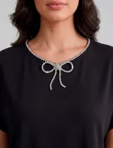 Blusa de modal suave color negro, con mangas cortas y corte holgado. Presenta un escote redondo decorado con una línea de strass y un broche desmontable en forma de moño con strass en el centro. La espalda cuenta con una abertura tipo gota con cierre de botón.
