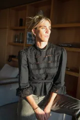 Blusa negra de manga 3/4 con cuello alto, confeccionada en algodón con paneles horizontales de encaje negro en el torso. Presenta un fajín integrado que se anuda a la cintura y puños con volado de encaje.