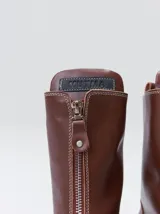 Botas de cuero color marrón de caña alta, con plataforma gruesa de goma y cierre metálico frontal con tirador. Presentan costuras a contratono y detalle de etiqueta de la marca en la lengüeta.