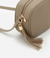 Cartera bandolera pequeña color beige, confeccionada en poliuretano con textura granulada. Presenta cierre superior con cremallera, correa larga ajustable y detalle de borla decorativa en el lateral con herrajes dorados.