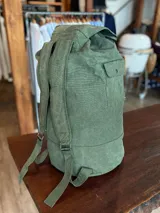 Mochila verde militar de lona con cierre superior con solapa y broche metálico. Tiene un bolsillo lateral con botón a presión y etiqueta de cuero con logo de la marca.