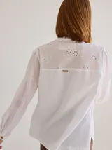 Camisa de corte recto y manga larga, confeccionada en algodón blanco. Presenta un canesú superior con bordado inglés floral y detalle de placa metálica con logo en la espalda.