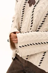 Buzo de tejido de punto color crudo con diseño de trenzas y costuras decorativas en color negro que recorren la prenda en forma de espiral. Presenta cuello redondo y mangas largas con terminación acampanada.