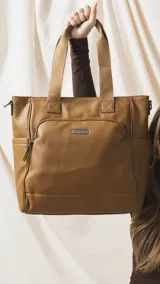 Cartera tipo tote marrón de diseño práctico y versátil, confeccionada en símil cuero con textura granulada. Cuenta con asas cortas fijas, bolsillos exteriores con cierre al frente y atrás, y bolsillos laterales abiertos. Su interior es amplio e incluye bolsillos organizadores. Viene con una correa ajustable y desmontable.