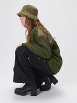 Campera tejido grueso y abrigado color verde militar, con forro de nylon interno, cuello estilo camisa en color negro y cierre frontal con botones. Cuenta con cuatro bolsillos delanteros con apliques en distintos tonos y texturas, y un parche bordado circular en el bolsillo superior izquierdo.