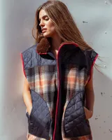 Chaleco sin mangas de diseño híbrido que combina paneles de tela acolchada capitoné en color azul marino con secciones de tejido de lana a cuadros en tonos naranja, gris y negro. Presenta cuello con forro de piel sintética marrón, cierre frontal con cremallera y ribetes en color rojo vibrante. Incluye bolsillos delanteros amplios con cierre.