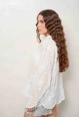 Camisa blanca de manga larga con bordados florales y detalles de ojales, cuello camisero y cierre frontal con botones ocultos.