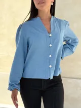 Camisa de jean celeste con cuello redondo, cierre frontal con botones y mangas largas abullonadas con puños ajustados.