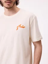 Remera de manga corta color crema con estampado gráfico en la espalda que representa un paisaje de montaña con un lago, árboles y una carpa, junto con el logo de la marca Rusty.