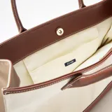 Bolso tipo tote de estructura rígida, confeccionado en lona color beige con detalles de cuero sintético en color marrón en la base, las asas y los bordes superiores.