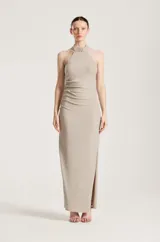 Vestido largo de fiesta color dorado con acabado brillante, escote halter y detalle de pedrería en el cuello. Presenta fruncidos en el lateral y una abertura en la falda.