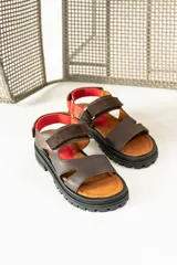 Sandalias de cuero marrón con diseño de tiras anchas y ajuste mediante velcro. Presentan una plantilla acolchada color tostado con detalles en rojo y una suela de goma negra con plataforma dentada.