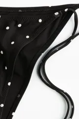 Bombacha de bikini vedetina negra con estampado de lunares blancos, tiro medio y tiras ajustables para atar en los laterales.