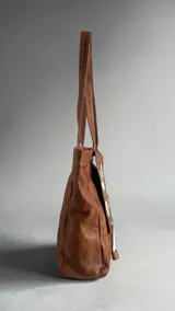 Cartera tipo tote de cuero color marrón terra, con diseño pintado a mano en el frente y borlas decorativas colgantes.