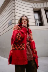 Saco tejido a mano con diseño jacquard en tonos rojo, naranja y marrón. Presenta cuello tipo polo, cierre frontal con botones grandes, mangas largas y tajos laterales en el ruedo.