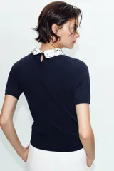 Jersey de punto negro de manga corta con cuello solapa blanco en contraste, decorado con abalorios joya y lentejuelas. Presenta cierre trasero con abertura y botón.