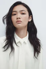 Camisa de manga larga con cuello clásico y botonadura frontal. Presenta una corbata extraíble a juego, puños con cierre de botón y un sutil detalle bordado en el pecho. El diseño incluye un canesú con pliegue en la espalda y un bajo redondeado con aberturas laterales.