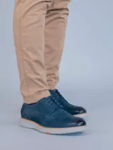 Zapato de estilo casual tipo derby en color azul marino, confeccionado en cuero con detalles de perforaciones decorativas tipo brogue. Cuenta con cordones al tono, detalle en cuero color suela en el talón y suela de goma flexible color gris claro con tecnología de amortiguación.