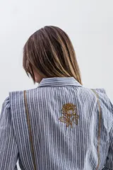 Camisa de algodón con estampado de rayas verticales en tonos gris y azulado. Presenta cuello clásico, cierre frontal con botones ocultos y detalles de pespuntes en hilo color óxido a lo largo del cuerpo y hombros. Incluye un bordado de rosa en la espalda.