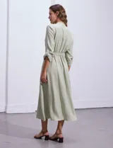 Vestido maxi color beige, con cuello mao y escote en V. Presenta mangas largas con presilla y botón para regular el largo, y un lazo ajustable en la cintura que marca la silueta.