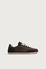 Zapatilla deportiva estilo barefoot, color marrón, con diseño de paneles en combinación de materiales y detalles en serraje. Cuenta con puntera redondeada y ancha, cordones en color crema y suela fina de goma flexible en contraste.