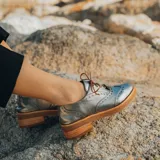 Zapato acordonado estilo oxford de cuero vacuno en color plateado metalizado, con diseño brogue, suela de goma y taco de madera.