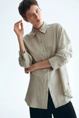 Camisa de corte oversized, color beige claro con rayas finas, cuello clásico, cierre frontal con botones, un bolsillo aplicado en el pecho, hombros caídos y mangas largas con puños abotonados.