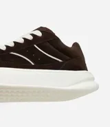 Championes de estilo skater para hombre, confeccionados en poliuretano color marrón oscuro. Poseen punta redonda, caña baja y suela de plataforma blanca. Destacan por sus cordones anchos tipo fat lace y detalles laterales en contraste.