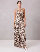 Vestido largo de tejido satinado plisado con estampado animal print tipo cebra en tonos tierra y ocre. Presenta escote halter profundo en V con drapeado central y broche de pedrería, espalda descubierta y falda fluida con apertura frontal.