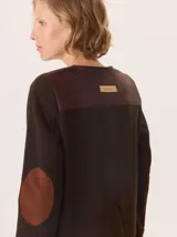 Chaqueta de paño color marrón oscuro, de corte clásico y cuello redondo, con cierre frontal dorado. Presenta bolsillos rectangulares con ribetes en color rojizo y parches de pana a rayas en los hombros.