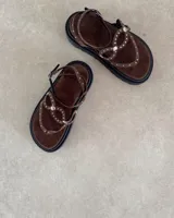 Sandalia de plataforma con base negra, confeccionada en gamuza marrón. Presenta tiras finas con apliques de tachas metálicas plateadas y cierre con hebilla ajustable en el tobillo.