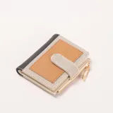 Billetera compacta de tres colores: negro en el borde, beige con patrón de puntos y un panel central marrón. Tiene un cierre de cremallera con un tirador en forma de imperdible dorado y un broche de solapa.