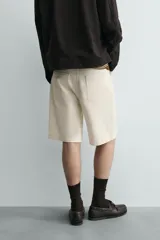 Bermuda de algodón color beige claro, de corte relajado y largo hasta la rodilla. Presenta cierre frontal con cremallera y botón, bolsillos delanteros y bolsillo cerillero, además de bolsillos traseros de plastrón.