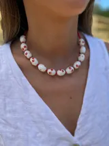 Collar corto con cuentas de cerámica blancas con estampado de flores rojas, separadas por mostacillas rojas. Cierre con cadena de acero quirúrgico y ganchito para regular el largo.