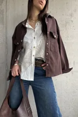 Camisa oversize blanca con botones frontales, cuello camisero y manga larga. Presenta grandes bordados en hilo beige con diseño de hojas o abanicos en la parte inferior frontal.