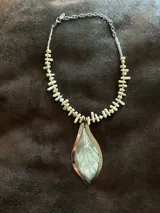 Collar corto con dije central en forma de hoja, elaborado en guampa con engarce de alpaca. La cadena está compuesta por bastoncitos de acrílico y mostacillas transparentes, finalizando con una cadena de eslabones de acero quirúrgico ajustable.