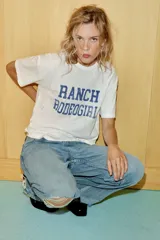Camiseta de corte oversize confeccionada en algodón flamé, con cuello redondo y manga corta. Presenta un diseño con efecto desgastado, detalles de roturas en el tejido y estampado de texto a contraste en la parte frontal.