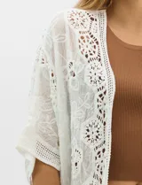 Cardigan abierto estilo kimono, color crudo, con mangas amplias al codo. Presenta una base con textura y motivos florales bordados a tono, con paneles de encaje calado estilo macramé en el frente, puños y ruedo.