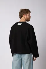 Buzo de cuello redondo confeccionado en tejido rústico con bordes al corte y costuras expuestas. Presenta un diseño boxy con estampado frontal y es reversible, permitiendo su uso por ambos lados.