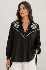 Camisa negra de manga larga con detalles bordados en contraste de color blanco en el pecho y hombros, cuello mao y cierre frontal parcial con botones.