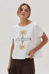Remera blanca de algodón con cuello bote y manga corta, con un estampado central que incluye dos palmeras reflejadas y la palabra "SUNRISE" bordada en el centro.