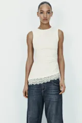 Camiseta ajustada (slim fit) color crema, con cuello redondo y mangas sisa. Presenta frunces laterales y un bajo asimétrico con detalle de encaje a tono.