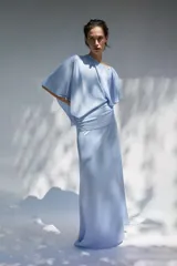 Vestido midi de tejido satinado color celeste, con cuello barco y diseño asimétrico con mangas tipo capa. Presenta un detalle drapeado en la cintura y abertura en la espalda.