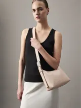 Cartera tipo hobo o baguette de piel sintética color rosa pálido, con correa de hombro ancha y ajustable. Presenta un diseño minimalista con el logo de Calvin Klein grabado sutilmente en el frente y herrajes metálicos plateados.